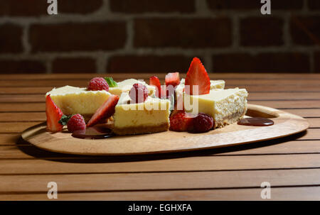 Il dessert. Torta di formaggio con lampone, fragola e salsa al cioccolato con foglia di menta su una lastra di legno. Muro di mattoni in background. Foto Stock