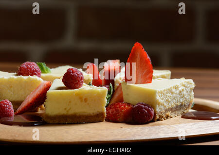 Il dessert. Torta di formaggio con lampone, fragola e salsa al cioccolato su un piatto di legno. Muro di mattoni in background. Foto Stock