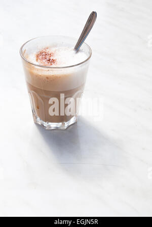 Cappuccino schiumoso in un caffè di vetro con cucchiaio su marmo bianco tabella all'aperto, dal lato. Foto Stock