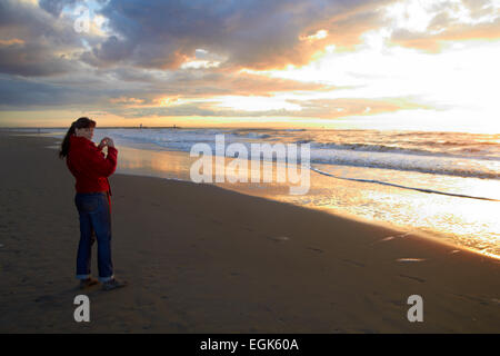 Donna di scattare una foto con il sole che tramonta dietro a freddo in spiaggia sabbiosa Foto Stock