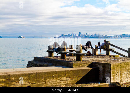 Un gruppo di giovani di relax sulle panchine di Sausalito in California con la skyline di San Francisco in distanza. Foto Stock