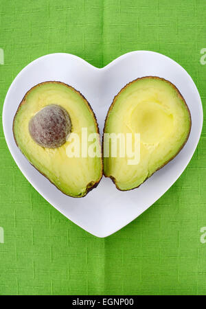 Avocado tagliato a metà a forma di cuore sulla piastra di colore verde brillante tovaglia, verticale. Foto Stock