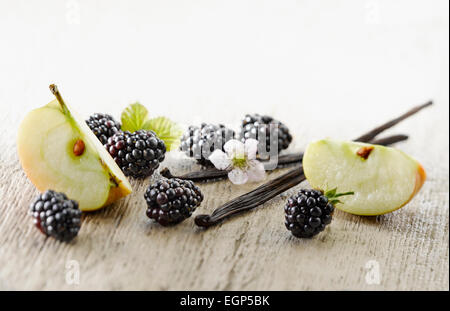 Blackberry, Rubus cultivar. Diverse bacche disposti con fiori e foglie e pezzi di mela, maggiorato di baccelli di vaniglia sul pallido, angosciato, sullo sfondo di legno. Messa a fuoco selettiva. Foto Stock