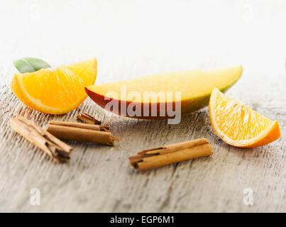 Mango, Mangifera indica. Uno strato disposto con quarti di Orange e pezzi rotti di stecca di cannella sul pallido, angosciato, sullo sfondo di legno. Messa a fuoco selettiva. Foto Stock