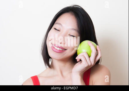 Giovane bella donna asiatica in abito rosso closeup sorridente con il chiudere gli occhi, mela verde sulla sua mano vicino a faccia su sfondo bianco Foto Stock