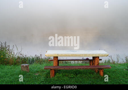 Foggy sunrize fiume. Panca e tavolo su fiume costa. Foto Stock