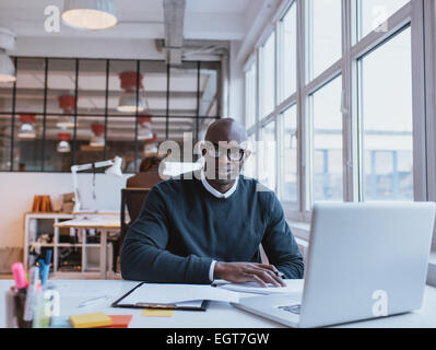 Ritratto di fiducioso giovane uomo seduto alla sua scrivania con il computer portatile. Imprenditore africani che lavorano in ufficio moderno. Foto Stock