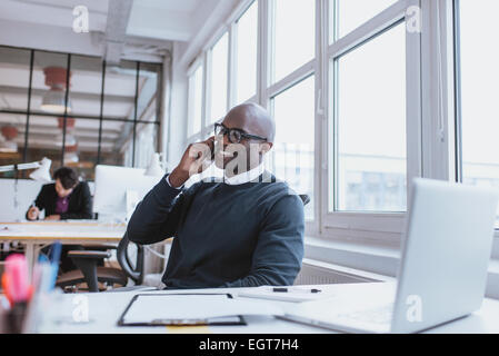 Giovane uomo che parla per il suo telefono cellulare in ufficio. African executive seduto alla sua scrivania con computer portatile Foto Stock