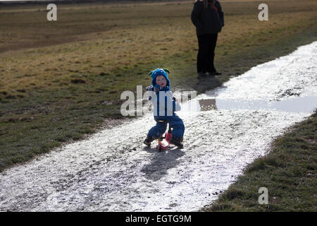 Un giovane bambino su uno scooter su un sentiero fangoso su un inverno pieno di sole il pomeriggio in sette sorelle Country Park. Foto Stock