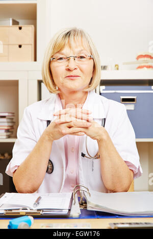 Vecchio sorridente medico donna seduta alla scrivania nel suo ufficio Foto Stock