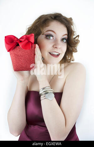Carino giovane donna che indossa senza spalline, borgogna abito rosso di contenimento, scintillanti confezione regalo con fiocco. Foto Stock