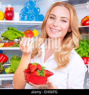 Closeup ritratto della bellissima femmina di mangiare fragole, aprire il frigo pieno di frutta e verdura, dieta e mangiare sano Foto Stock