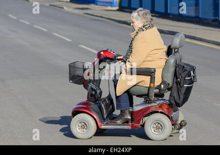 Mobilità scooter - signora anziana attraversare una strada in una mobilità scooter NEL REGNO UNITO. Senior donna disabilità scooter. Foto Stock