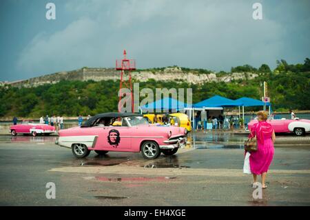 Cuba, La Habana, donna in abito rosa attraversando la strada con molti vecchi american chevrolet parcheggiata Foto Stock