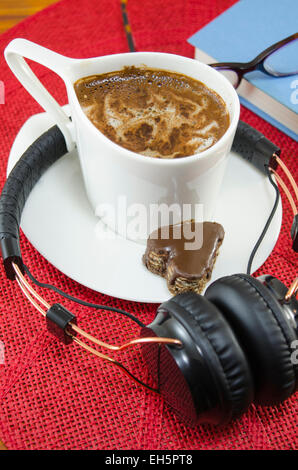 Tazza di caffè e cuffie, libro e occhiali su una tovaglia rosso Foto Stock