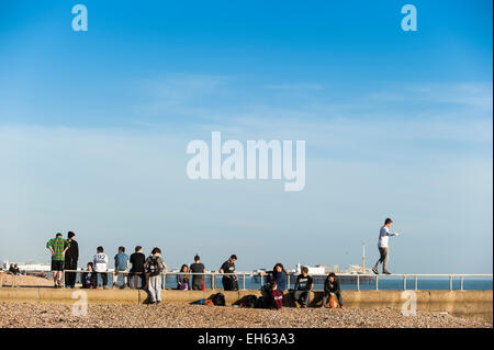 Un giovane uomo cammina in equilibrio su un elevato una rampa mentre un gruppo di giovani che stanno facendo la maggior parte della soleggiata primavera meteo praticare l'arte del parkour, free-jumping e facendo salti mortali, off un pontile sul lungomare di Brighton con il dis-usato Molo Ovest in background. Brighton, East Sussex, Inghilterra, Regno Unito. Foto Stock