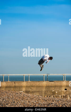 Un giovane uomo di torsioni in aria mentre un gruppo di giovani che stanno facendo la maggior parte della soleggiata primavera meteo praticare l'arte del parkour, free-jumping e facendo salti mortali, off un pontile sul lungomare di Brighton con il dis-usato Molo Ovest in background. Brighton, East Sussex, Inghilterra, Regno Unito. Foto Stock