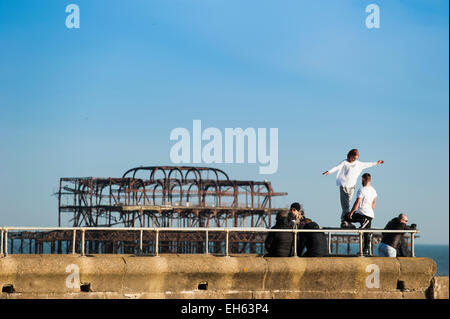 Un hipster uomo saldi in una posizione sollevata della rampa di metallo mentre un gruppo di giovani che stanno facendo la maggior parte della soleggiata primavera meteo praticare l'arte del parkour, free-jumping e facendo salti mortali, off un pontile sul lungomare di Brighton con il dis-usato Molo Ovest in background. Brighton, East Sussex, Inghilterra, Regno Unito. Foto Stock