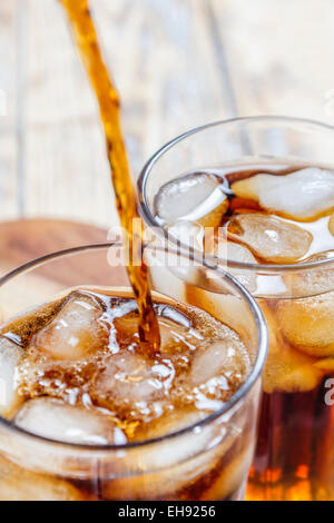 Cola versando in un bicchiere con ghiaccio a cubetti con un altro vetro già riempito su tavola in legno rustico Foto Stock