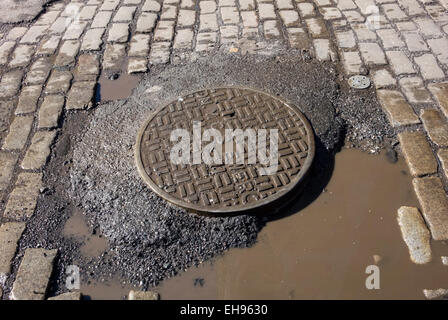 Tombino di fognature e coperchio blocco belga acciottolato in necessità di riparazione sulla strada di SoHo a New York City Foto Stock