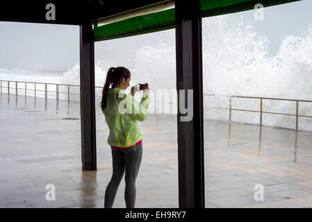 Le donne di fotografare grandi onde che si infrangono aginst broadwalk con telefono cellulare, vestito in marcia Foto Stock