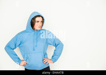 Un moody 13 14 15 anno di età giovane ragazza adolescente che indossa un blu felpa con cappuccio Felpa con cappuccio Felpa in piedi contro un bianco di sfondo per studio REGNO UNITO Foto Stock