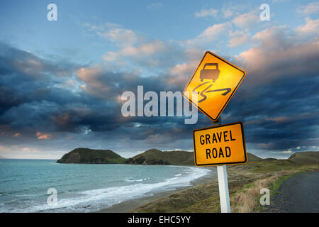 Attenzione (strada di ghiaia) segno sulla strada per mare contro il cielo nuvoloso al tramonto. Foto Stock