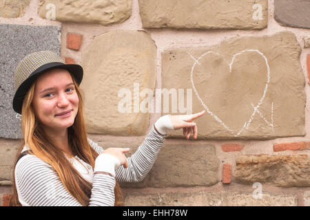 Giovane ragazza carina nel cappello vicino al muro con disegnate chalk cuore. Foto Stock