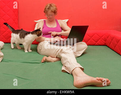 Donna con notebook seduta sul suo letto e il suo gatto accanto a lei Foto Stock