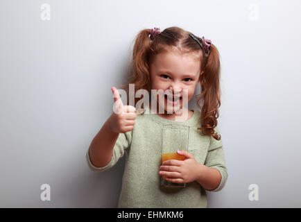 Ridendo kid ragazza di bere succo di frutta e mostrando pollice in alto segno su sfondo blu Foto Stock