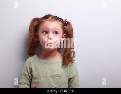 Divertente pensare kid ragazza mangiare lecca-lecca e guardando in alto su sfondo grigio Foto Stock