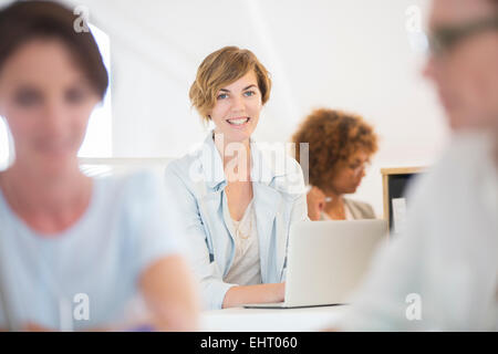 Ritratto di donna seduta alla scrivania, utilizzando laptop e sorridente in ufficio, i colleghi in background Foto Stock