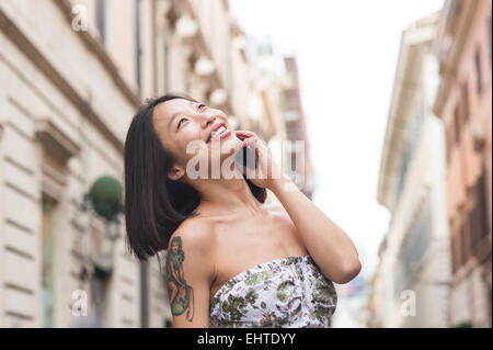 Giovane bella donna asiatica con tatuaggio sulla spalla e sorridente a parlare utilizzando il telefono cellulare all'aperto urbano Foto Stock