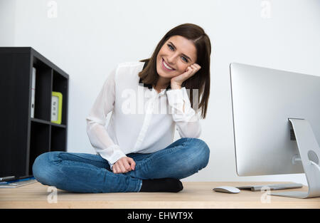 Sorridente giovane imprenditrice seduta sul tavolo in ufficio Foto Stock