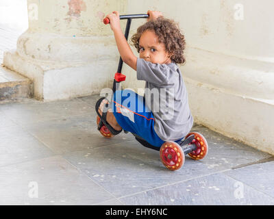 Un cubano toddler maschio con ricci capelli castani si siede sul suo scooter a tre ruote e guarda oltre la sua spalla alla fotocamera. Foto Stock