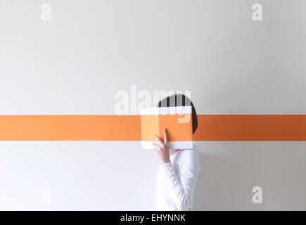 Vista laterale di una donna che nasconde il suo volto dietro un libro con una striscia arancione Foto Stock