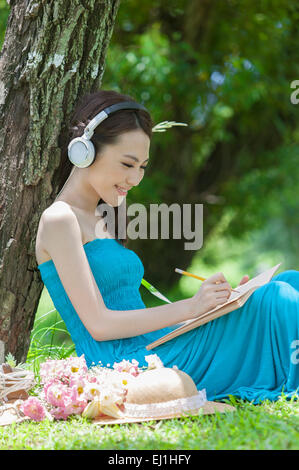 Giovane donna seduta sul prato, indossando le cuffie e scrivendo con il sorriso, Foto Stock