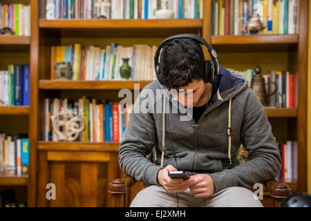 Attraente giovane uomo seduto per ascoltare musica su un set di cuffie stereo nel suo studio di fronte a una libreria riempito con bo Foto Stock