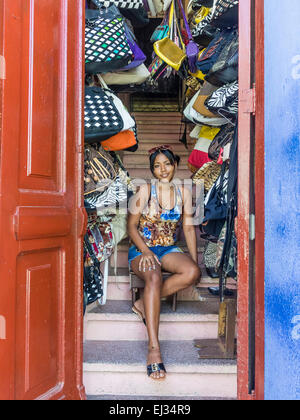 Una femmina ispanica negoziante siede su scale nell'apertura porta del suo piccolo negozio che vende portamonete in Santiago de Cuba Foto Stock