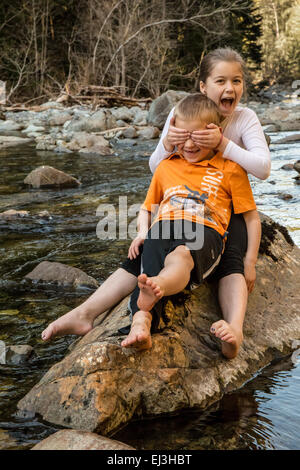 9 anno vecchia ragazza scherzosamente che copre gli occhi dei suoi sette anni di fratello come loro siedono su un masso in un fiume poco profondo Foto Stock