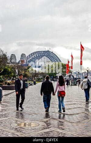 Persone non identificate a piedi lungo una banchina umido a Sydney in Australia dopo un temporale con il Ponte del Porto di Sydney in anticipo. Foto Stock