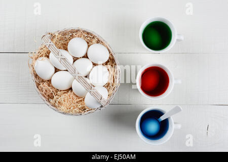 Alta angolazione di un cesto pieno di uova per essere tinti per la Pasqua. Tre tazze di colorante con uova ammollo sono accanto ai cestelli di u Foto Stock