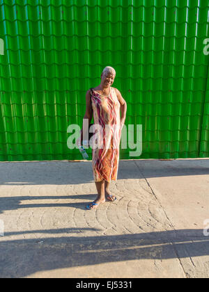 Un afro-cubane senior citizen donna con i capelli grigi si erge contro un verde Sfondo piastrelle che indossa un abito lungo e rivolto in avanti Foto Stock