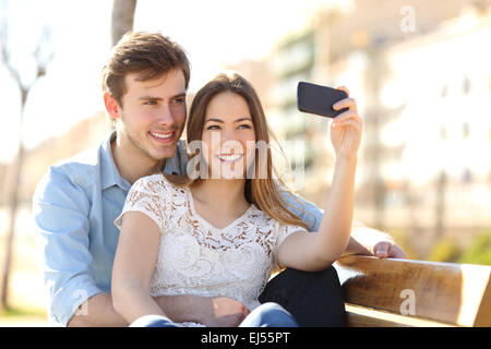 Giovane fotografa un selfie con uno smart phone in un parco con un background urbano in una giornata di sole Foto Stock