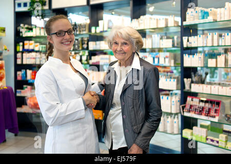 Sorridente e simpatico farmacista femmina che indossano occhiali assistere una signora anziana nella sua farmacia o farmacia agitando mani con l Foto Stock