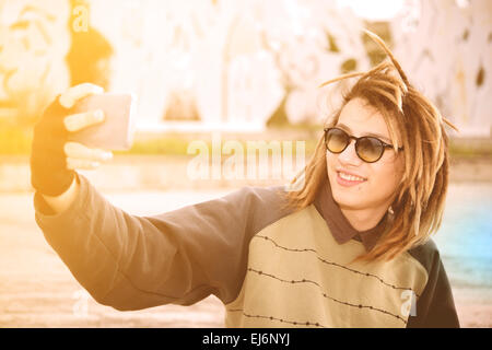 Ritratto di giovane ragazzo outdoor con capelli rasta sorridente con smart phone in un concetto di stile di vita con un caldo filtro applicato Foto Stock