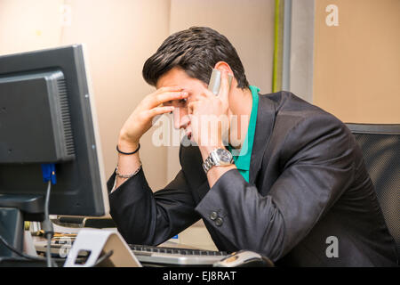 Grave attraente giovane imprenditore seduto alla scrivania in ufficio occupato parlando al telefono Foto Stock
