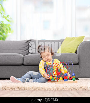 Carino bambina gioca con un giocattolo seduto sul pavimento accanto al grigio di un divano a casa. Foto Stock