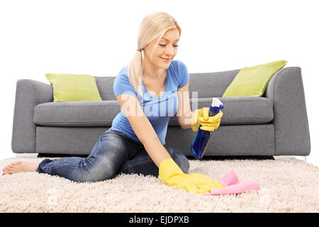 Donna di pulizia di un tappeto con uno spray di pulizia e giallo da indossare guanti di protezione Foto Stock