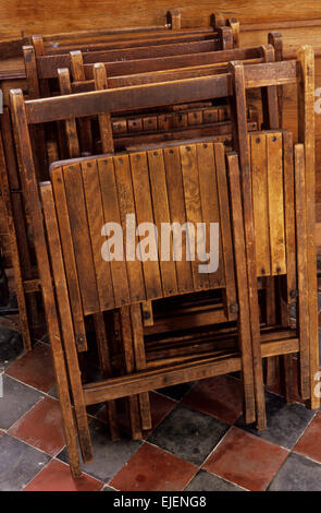 Vintage marrone pieghevoli sedie di legno sovrapposti in fila contro la parete e in piedi sul nero e piastrelle color ocra Foto Stock